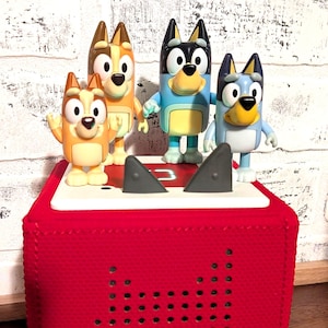 May include: A red audio player with a speaker design and a set of four cartoon dog figurines. The figurines are in various poses and colors, including blue, orange, and black. The background is a white brick wall.