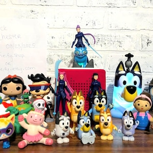 May include: A collection of colorful figurines, including characters from popular animated series and movies. The figurines are arranged on a wooden surface against a white brick wall. Various sizes and styles are present.