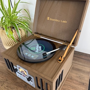 May include: A wooden Bambu Lab record player with a built-in screen. The record player has a dark brown finish with a light brown top and a small orange knob. The screen displays a graphic with the Bambu Lab logo. The record player is on a wooden surface.