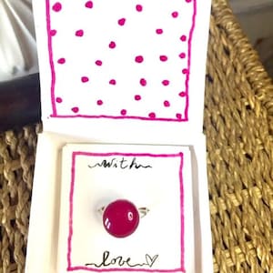 Può includere: Una confezione regalo fatta a mano con un anello all'interno. L'anello presenta una grande pietra rotonda color magenta montata su una fascia argentata. La scatola è bianca con un bordo rosa e le parole "with love".