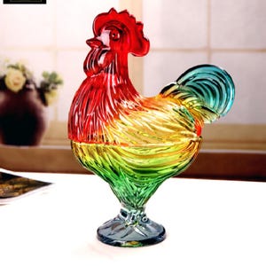 May include: A colorful glass rooster figurine. The rooster features a gradient of colors, starting with red at the comb and transitioning to yellow, green, and blue in the tail. The figurine is on a clear, pedestal base.