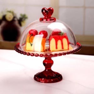 Può includere: Un alzatina per torte in vetro rosso con una cupola in vetro trasparente. Il supporto ha una maniglia a forma di cuore e un bordo perlato. Sotto la cupola sono visibili due fette di torta, guarnite con ciliegie e fragole.