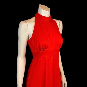 May include: A bright red halter-neck dress. The dress has a high neckline, a fitted bodice, and a flowing skirt. The dress is displayed on a mannequin against a black background. The dress is a solid colour and appears to be made of a smooth fabric.