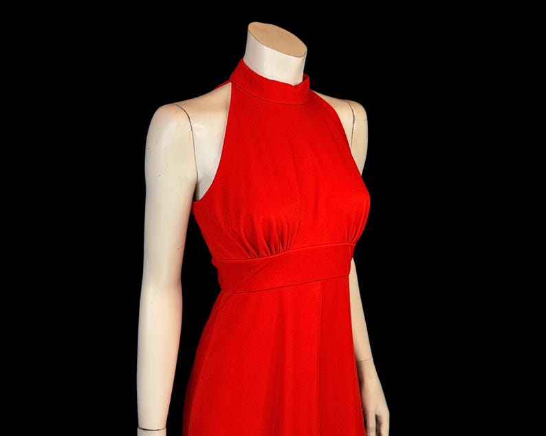 May include: A bright red halter-neck dress. The dress has a high neckline, a fitted bodice, and a flowing skirt. The dress is displayed on a mannequin against a black background. The dress is a solid colour and appears to be made of a smooth fabric.