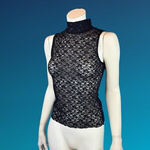 May include: A black sleeveless top made of delicate lace. The top features a high, mock neck and a floral pattern. The garment is displayed on a mannequin against a blue background. The top is a stylish piece of apparel.