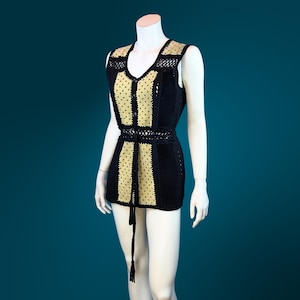 May include: A black and gold crochet vest with a V-neck design. The vest features a fitted silhouette with a tie at the waist and a decorative fringe. The gold sections have a dotted pattern. The vest is displayed on a mannequin.
