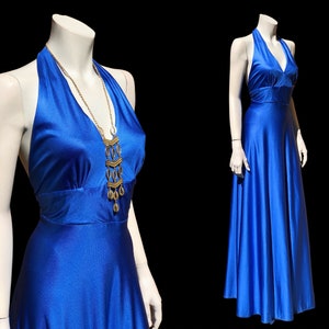 May include: A royal blue halter-style maxi dress with a V-neckline and a flowing skirt. The dress is accessorized with a gold chain necklace with a pendant featuring multiple circular and oval-shaped charms.