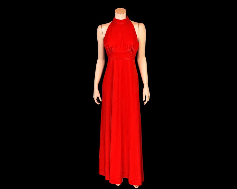 May include: A striking red halter-neck evening gown. The dress has a fitted bodice, a cinched waist, and a flowing, floor-length skirt. The gown is displayed on a mannequin against a black backdrop, emphasizing its elegant design.