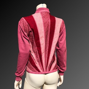 May include: A burgundy zip-up jacket with a unique design. The back features a sunburst pattern in varying shades of red, with textured and patterned panels. The sleeves and collar are a solid burgundy color, creating a striking contrast.