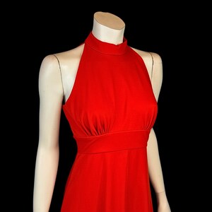 May include: A bright red halter dress on a mannequin. The dress has a high neckline, fitted bodice, and a flowing skirt. The fabric looks smooth and has a slight sheen. The dress is a solid colour, with no visible patterns or embellishments.