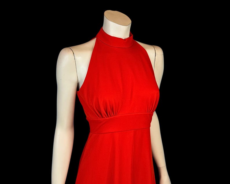 May include: A bright red halter dress on a mannequin. The dress has a high neckline, fitted bodice, and a flowing skirt. The fabric looks smooth and has a slight sheen. The dress is a solid colour, with no visible patterns or embellishments.