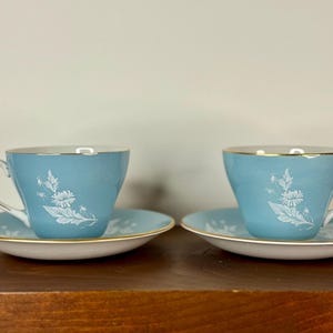 May include: Two matching blue teacups and saucers with a white floral design and gold trim. The teacups have a curved handle and sit on a white saucer with a gold rim. The teacups and saucers are displayed on a wooden surface.