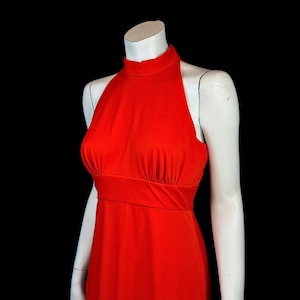 May include: A bright red halter dress on a white mannequin against a black backdrop. The dress has a high neckline, fitted bodice, and a flowing skirt. The fabric looks like a smooth, stretchy material.