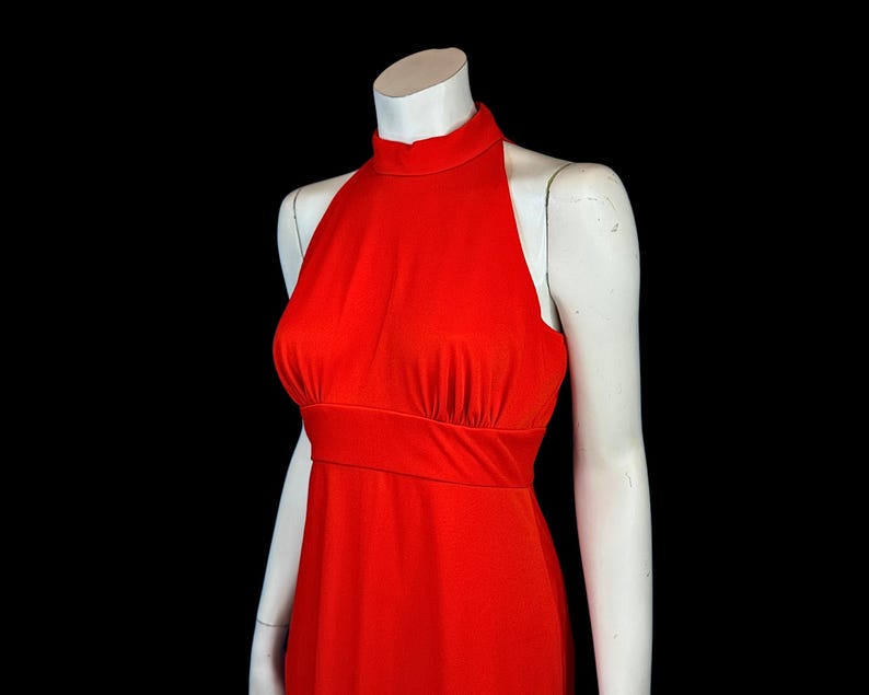 May include: A bright red halter dress on a white mannequin against a black backdrop. The dress has a high neckline, fitted bodice, and a flowing skirt. The fabric looks like a smooth, stretchy material.