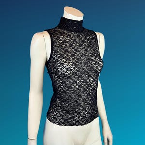May include: A black lace sleeveless top with a high neckline is displayed on a mannequin. The intricate floral lace pattern covers the entire top, creating a delicate and see-through effect. The mannequin is set against a blue background.