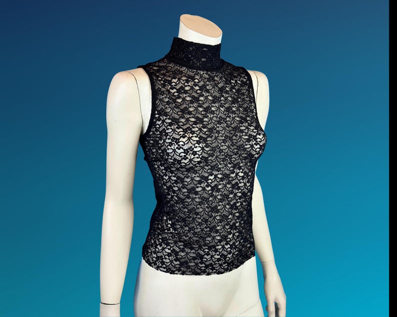 May include: A black lace sleeveless top with a high neckline is displayed on a mannequin. The intricate floral lace pattern covers the entire top, creating a delicate and see-through effect. The mannequin is set against a blue background.