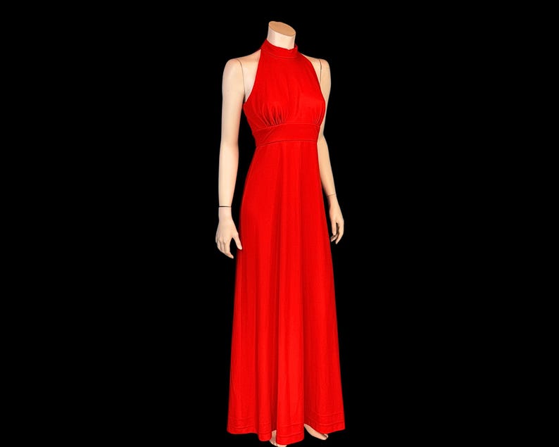 May include: A bright red halter-neck maxi dress. The dress has a fitted bodice and a flowing skirt. The dress is displayed on a mannequin against a black background. The dress is a solid colour and appears to be made of a soft fabric.
