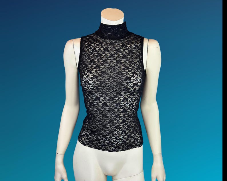 May include: Black sleeveless lace top with a mock neck. The intricate floral lace pattern covers the entire top. The top is displayed on a mannequin against a blue background.