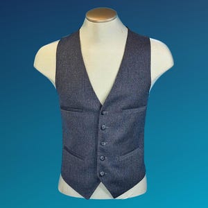 May include: A dark grey waistcoat featuring a subtle vertical pinstripe pattern, a V-neckline, and five matching buttons down the centre. The garment includes two horizontal welt pockets on the front and a pointed hemline at the bottom.