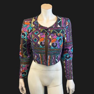 May include: A cropped long-sleeve jacket featuring a vibrant patchwork pattern with geometric shapes and abstract motifs in shades of purple, teal, orange, and black. The jacket has a round neckline, a single front closure, and long sleeves.