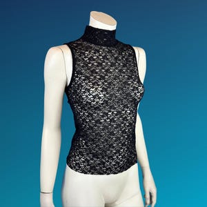 May include: Black sleeveless lace top with a mock neck. The top is displayed on a mannequin against a blue background. The intricate floral lace pattern covers the entire garment, creating a delicate and elegant look.