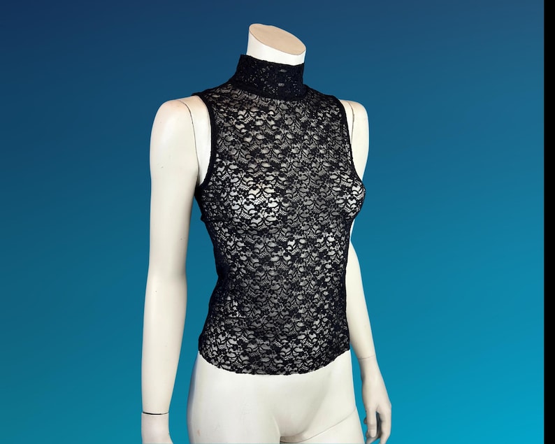 May include: Black sleeveless lace top with a mock neck. The top is displayed on a mannequin against a blue background. The intricate floral lace pattern covers the entire garment, creating a delicate and elegant look.