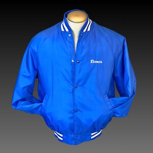 May include: A royal blue satin baseball jacket with white ribbed trim around the collar, cuffs, and waistband. The jacket has a button closure and a white embroidered name "Barnes" on the left chest.