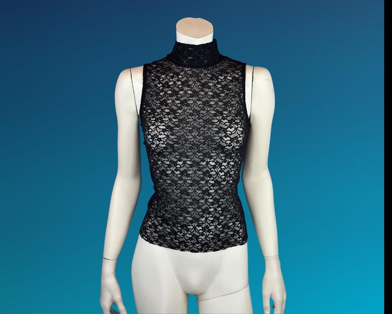 May include: Black sleeveless lace top with a mock neck. The top is displayed on a mannequin against a blue background. The lace pattern is intricate and covers the entire garment.