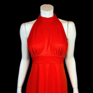 May include: A bright red halter dress with a high neckline and a fitted waist. The dress is displayed on a white mannequin against a black background, highlighting its sleek design and bold colour. The dress is a solid colour and appears to be made of a smooth fabric.