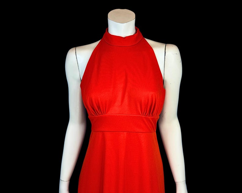 May include: A bright red halter dress with a high neckline and a fitted waist. The dress is displayed on a white mannequin against a black background, highlighting its sleek design and bold colour. The dress is a solid colour and appears to be made of a smooth fabric.