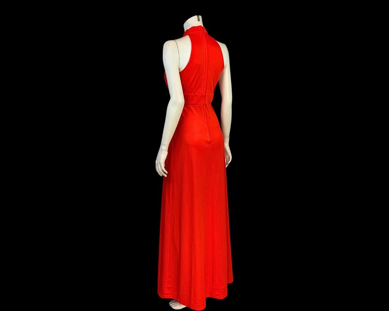 May include: A striking red, full-length dress with a high neck and no sleeves. The gown has a fitted bodice and a flowing skirt, creating an elegant silhouette. The dress is displayed on a mannequin against a black background.