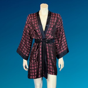 May include: A black and fuchsia-colored kimono-style robe with a repeating pattern. The robe has a black satin collar, cuffs, and belt. The belt is tied at the waist. The robe is displayed on a mannequin against a blue background.