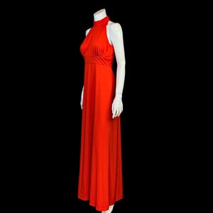 May include: A striking, full-length, halter-neck dress in a vivid red hue. The dress has a fitted bodice and a flowing skirt, creating an elegant shape. The dress is displayed on a white mannequin against a black backdrop.
