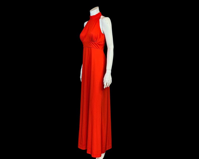 May include: A striking, full-length, halter-neck dress in a vivid red hue. The dress has a fitted bodice and a flowing skirt, creating an elegant shape. The dress is displayed on a white mannequin against a black backdrop.