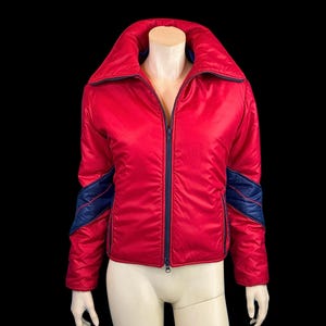 May include: A red puffer jacket with blue accents on the sleeves and collar. The jacket has a zip closure and a stand-up collar.