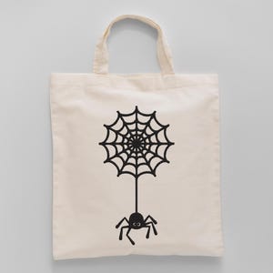 K&ouml;nnte beinhalten: Eine cremefarbene Tragetasche mit einem schwarzen Spinnennetz-Design und einer darunter h&auml;ngenden Spinne. Die Tasche hat zwei Griffe und besteht aus einem hellfarbigen Stoff, ideal zum Tragen von Gegenst&auml;nden.