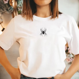 K&ouml;nnte beinhalten: Wei&szlig;es T-Shirt mit einem schwarzen Spinnenmuster auf der Brust. Das kurz&auml;rmelige Oberteil ist aus einem weichen Stoff gefertigt. Die Spinnenillustration ist ein einfaches, stilisiertes Design.