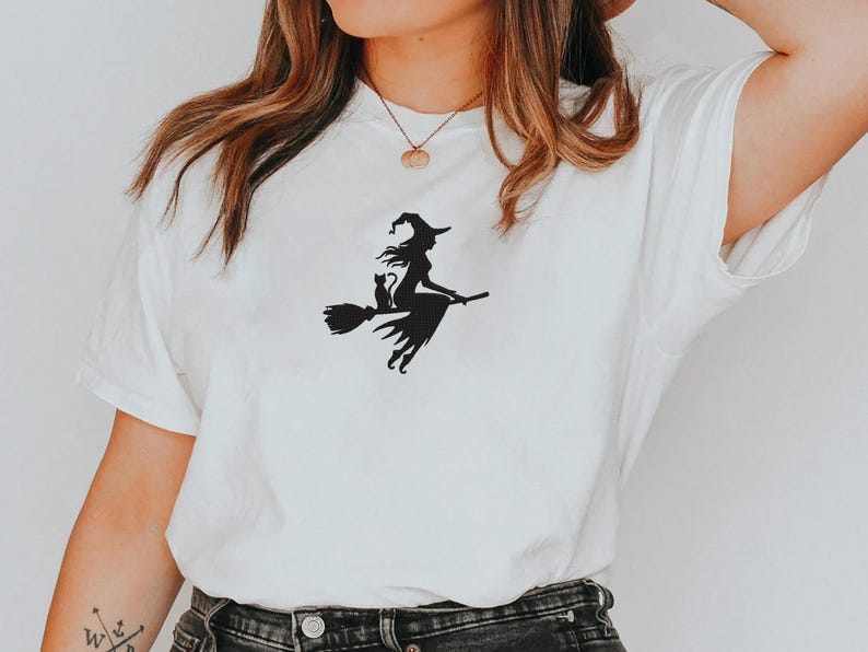 May include: White t-shirt featuring a black silhouette of a witch riding a broom with a black cat. The witch is wearing a pointed hat and flowing dress. The design is centered on the front of the shirt.