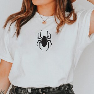 K&ouml;nnte beinhalten: Wei&szlig;es T-Shirt mit einem schwarzen Spinnenmuster. Die Spinne ist mittig platziert und hat ein detailliertes, symmetrisches Design. Das Shirt besteht aus einem weichen Material und hat kurze &Auml;rmel.
