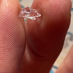 May include: A small, clear, crystalline object of irregular shape is held between two fingers. The object is translucent and reflects light, possibly a gemstone or decorative element.