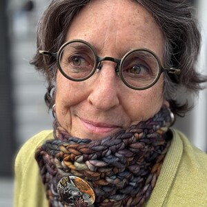May include: A thick, hand-knitted cowl in a mix of brown, grey, and purple yarns, fastened with a decorative button. The button has a nature-inspired design. The cowl is worn over a yellow cardigan.