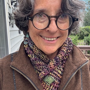 May include: A hand-knitted cowl in variegated shades of purple, yellow, and brown, secured with a green ginkgo leaf-shaped pin. The cowl is worn over a brown zip-up jacket, paired with round-framed glasses and dangling earrings.