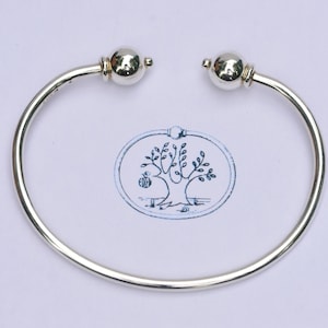 May include: A polished silver cuff bracelet featuring a smooth, open-ended design with two spherical ball terminals at each end. The bracelet has a reflective metallic finish and a circular shape, showcasing a clean and simple aesthetic.