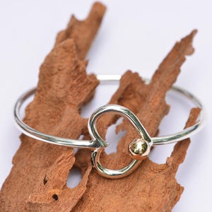 May include: A silver bangle bracelet featuring a central heart-shaped design element. The heart is accented with a small, polished gold-toned sphere positioned at the lower right curve of the heart shape. The bracelet has a smooth, reflective finish.