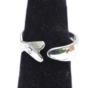 May include: A silver-toned open ring featuring a fish design. The band is shaped like a fish body with a detailed tail fin on one end and a smooth head with a small gold-colored accent on the eye on the other end, creating a wrap-around effect.