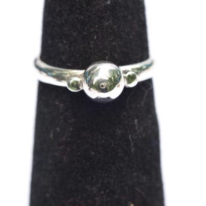 May include: A polished silver ring featuring a central spherical bead flanked by two smaller spherical beads on either side. The band is smooth and metallic, showcasing a reflective surface that catches the light, creating a clean and simple design.