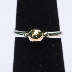 May include: A two-tone ring featuring a polished silver band with a central gold-colored sphere. The design includes a smooth, rounded metal finish on the band and a prominent, reflective gold ball set at the center of the ring's front surface.