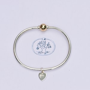 May include: A silver bangle bracelet featuring a single polished gold-tone bead at the top and a small heart-shaped charm dangling from the bottom. The bracelet is centered over a blue ink stamp illustration of a tree inside an oval border.