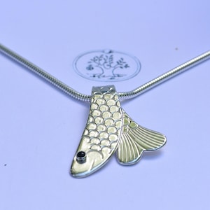 May include: A silver-toned metal necklace featuring a pendant shaped like a fish with a textured scale pattern on its body, a small black circular eye, and a fanned tail. The pendant hangs from a smooth, round silver-toned snake chain necklace.