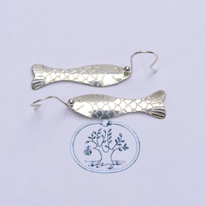May include: A pair of silver-toned fish-shaped earrings with engraved scale patterns and curved ear wires. Below the earrings is a circular stamp featuring a line drawing of a tree with the word EDEN written on the trunk.